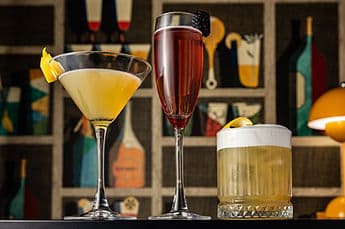 Three signature cocktails on the bar against a painted-bottle backdrop.